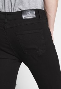 Vaqueros de mezclilla negros con un bolsillo de parche acolchado y una etiqueta de cuero en la cintura con el logotipo de mariposa. Silueta ajustada con una ligera textura.