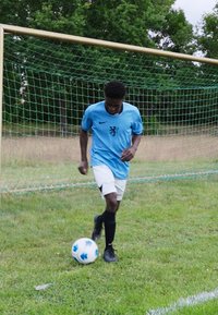 Un giocatore in una maglia Nike azzurro chiaro con un logo di leone, pantaloni corti bianchi e calze nere, dribbla un pallone da calcio su un terreno erboso.
