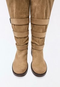Bottes hautes en daim brun clair avec trois larges sangles horizontales et des semelles plates marron foncé sur fond blanc.
