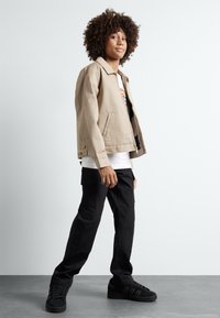 Beige jacket with a collar, black cargo pants, and black sneakers. Model stands sideways with a textured curly hairstyle.