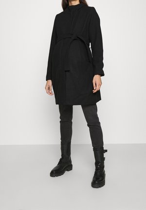 Femme portant un manteau noir ceinturé, un jean skinny foncé et des bottes noires à lacets, debout devant un fond uni clair.