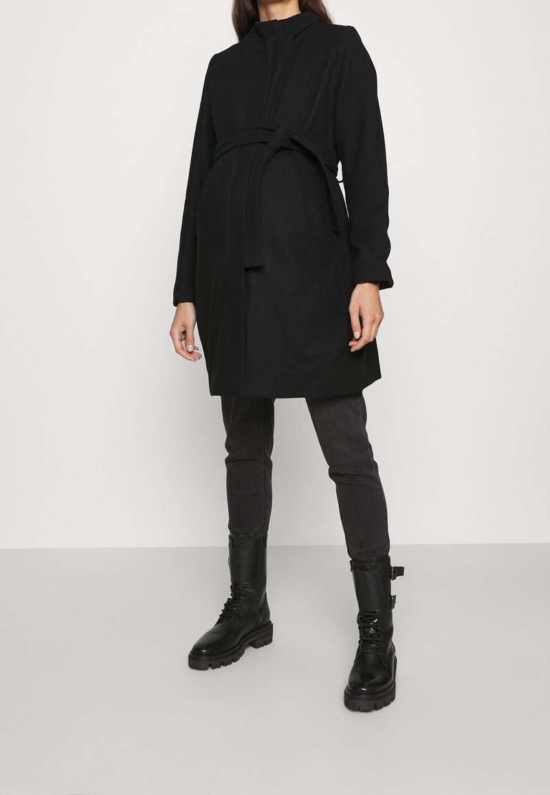 Femme portant un manteau noir ceinturé, un jean skinny foncé et des bottes noires à lacets, debout devant un fond uni clair.
