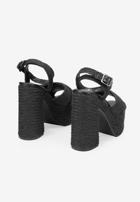 Sandalias negras de tacón alto con textura trenzada, correa en el tobillo con hebilla y tacones gruesos estilo cuerda. Plantilla suave con recortes redondeados.