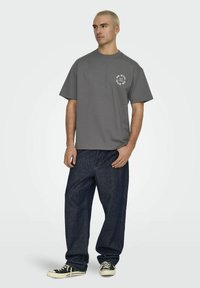 Graues Baumwoll-T-Shirt mit rundem Halsausschnitt und kurzen Ärmeln; versehen mit einem weißen kreisförmigen Grafikdesign auf der Brust. Kombiniert mit dunkelblauen Jeans.