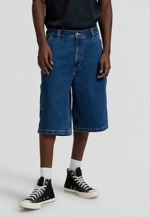 Pantalones cortos de mezclilla en azul oscuro, hasta la rodilla, con un diseño clásico de cinco bolsillos y costuras en contraste. Combinados con zapatillas negras de caña alta.