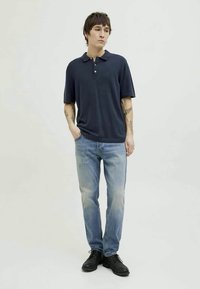 Νεαρός άνδρας με κοντά σκούρα μαλλιά, φορώντας ναυτικό μπλουζάκι, ανοιχτόχρωμα τζιν, μαύρα παπούτσια και τατουάζ στα δύο μπράτσα, στέκεται με τα χέρια στις τσέπες.