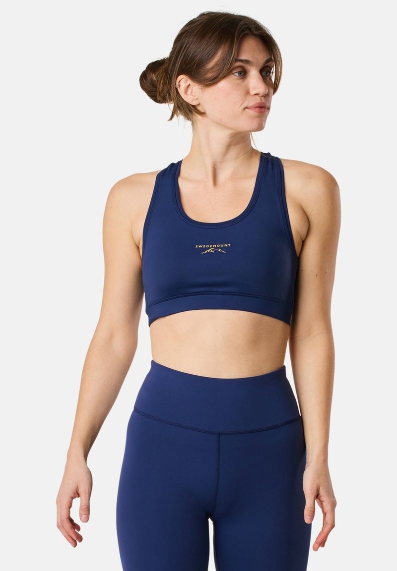 Granatowy stanik sportowy z zaokrąglonym dekoltem, wycięciem z tyłu, miękką teksturą oraz małym złotym logo na środku. Odpowiedni do odzieży sportowej.