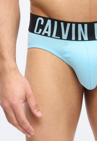 Ανδρικός κορμός και χέρι που φορούν ανοιχτό μπλε εσώρουχο με μαύρη μέση και μεγάλο κείμενο "Calvin".