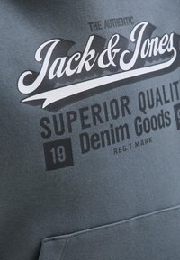 Artimas vaizdas, kuriame matomas pilkas audinys su baltu ir juodu tekstu: "Autentiškas Jack & Jones Aukščiausios kokybės 19 džinsų prekės ženklas."