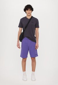 Purple shorts and a dark gray t-shirt are worn with white sneakers. A small black crossbody bag is slung across the shoulder.