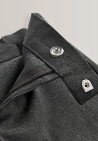 Gros plan sur la ceinture d'un pantalon gris foncé avec un bouton transparent, une agrafe en métal et une fermeture éclair partiellement visible.