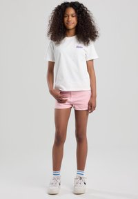 White cotton t-shirt with purple logo, paired with light pink denim shorts. White sneakers with black accents and blue-striped socks.