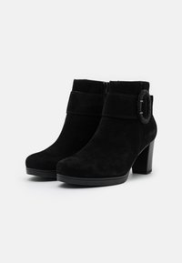 Botas tobilleras de ante negro con tacón alto, cierre de cremallera y un acento redondo decorativo en la correa del tobillo. Textura suave en todo.