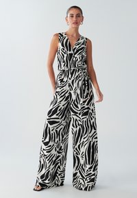 Zwart-witte dierenprint jumpsuit met een diepe V-hals, taille die is aangelijnd, en een broekspijp met een wijd ontwerp. De stof lijkt lichtgewicht en vloeiend.