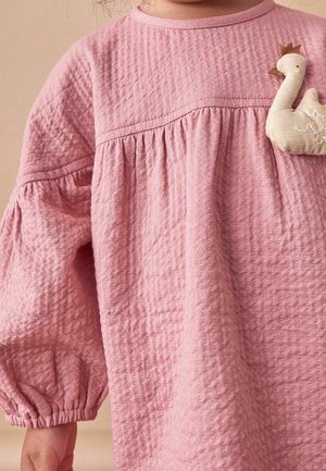 Enfant portant une robe rose texturée à manches longues avec poignets élastiques et une broche en tissu beige en forme de cygne épinglée près de l'épaule.