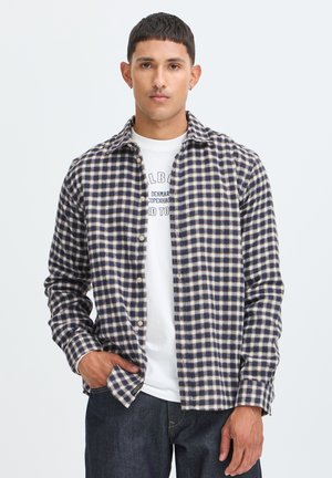 Jonge man die een zwart-wit geruit overhemd draagt over een wit grafisch T-shirt en donkere jeans, staand met één hand in zijn zak.