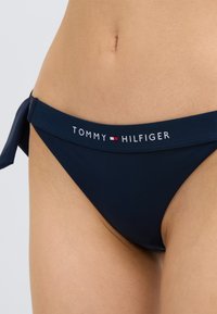 Μπλε σκούρο μαγιό μπικίνι με ρυθμιζόμενο δετό πλαϊνό, με λευκή λωρίδα λογότυπου που γράφει "TOMMY HILFIGER" και κόκκινες και λευκές λεπτομέρειες.