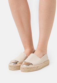 Beige espadrilles dengan kap toe emas berkilau, platform yang dibalut jute, dan bagian atas kulit halus dengan desain belakang terbuka.