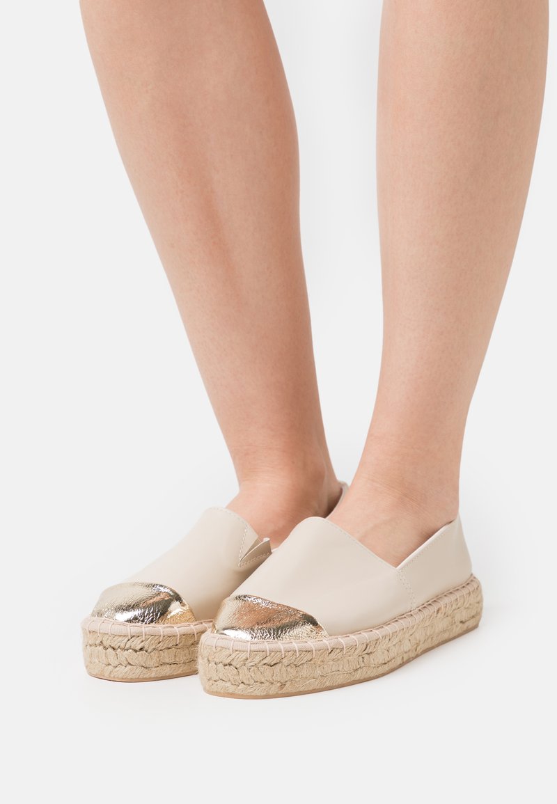 Beige espadrilles dengan kap toe emas berkilau, platform yang dibalut jute, dan bagian atas kulit halus dengan desain belakang terbuka.