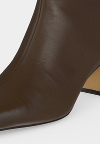 Gros plan d'une botte en cuir marron à bout pointu avec couture cousue et talon bloc visible sur fond blanc.