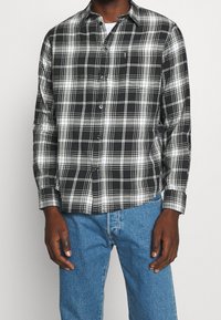 Camicia a quadri bianca e nera con colletto, maniche lunghe e chiusura frontale a bottoni. Indossata sopra una maglietta bianca, abbinata a jeans blu.