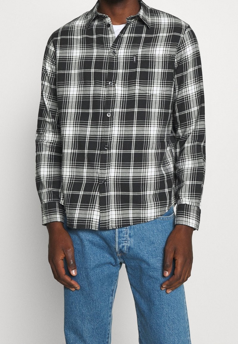 Camicia a quadri bianca e nera con colletto, maniche lunghe e chiusura frontale a bottoni. Indossata sopra una maglietta bianca, abbinata a jeans blu.