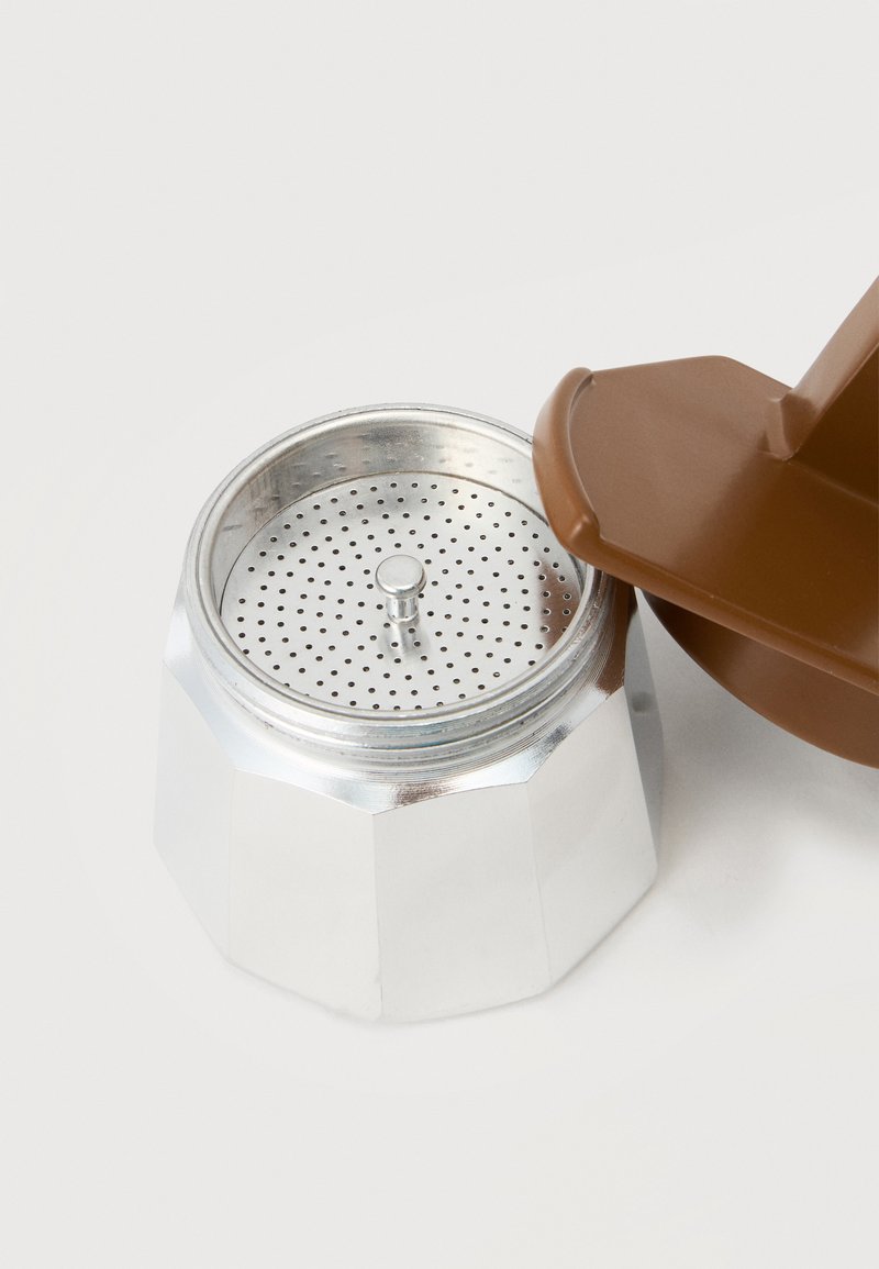 Aluminium Herd Espressomaschinenbasis mit perforiertem Filter und braunem Kunststofftrichterdeckel daneben auf weißer Oberfläche.
