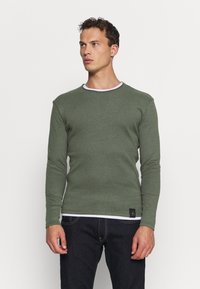 Grüner Pullover mit langen Ärmeln und weißem Kragen, strukturierter Stoff, figurbetonte Passform und einem kleinen Logopatch am Saum. Getragen mit dunklen Jeans.