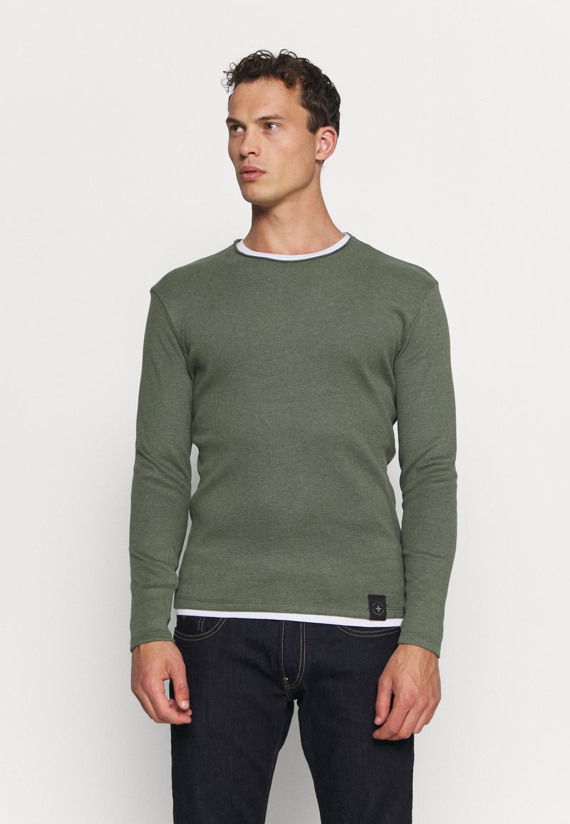 Grüner Pullover mit langen Ärmeln und weißem Kragen, strukturierter Stoff, figurbetonte Passform und einem kleinen Logopatch am Saum. Getragen mit dunklen Jeans.