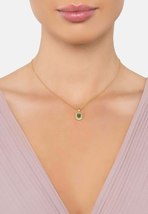 Collar de cadena de oro con colgante rectangular de piedra preciosa verde rodeado de pequeñas piedras claras, llevado por una mujer con un top de cuello en V de color púrpura claro.