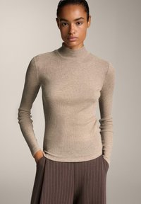 Kvinde iført en tætsiddende beige ribbet turtleneck sweater og brune stribede bukser med brede ben, med hænderne i lommerne, neutral baggrund.