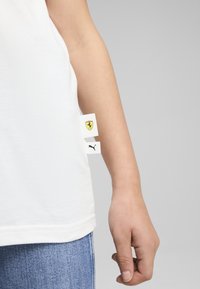 Valkoinen puuvillapaita, jossa on logo-tagit Ferrarilta ja Pumalta sivussa. Suora helma, minimalistinen muotoilu, pehmeä tekstuuri näkyvissä.