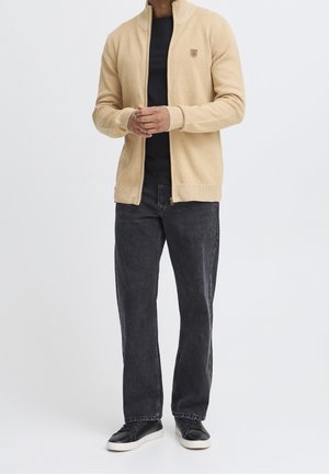 Beige neulevetoketjutakki, jossa korkea kaulus ja pieni logo rinnassa. Päällä musta paita, tummat farkut ja lenkkarit.