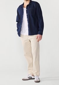 Denimskjorta i mörkblått, knapplåst design med en bröstficka, kombinerad med beige byxor och vita sneakers med svarta ränder.