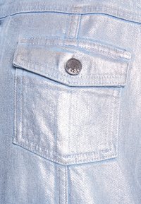 Giacca di jeans azzurro chiaro con finitura metallica, dotata di tasca sul petto e dettagli in bottoni a pressione argentati. Texture liscia e design strutturato.