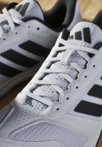 Des chaussures de sport blanches avec des accents noirs, en matériau synthétique et en maille, dotées d'un système de laçage proéminent et de détails perforés sur l'orteil.