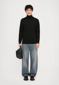 Hombre con cabello rizado, vistiendo un jersey de cuello alto negro, vaqueros anchos azul desgastado, zapatos oscuros, sosteniendo un bolso de cuero negro frente a un fondo blanco.