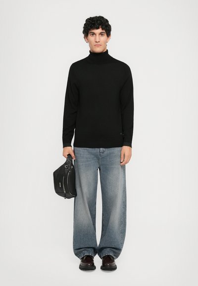 Mann med krøllete hår iført svart høyhalset genser, vide falmede blå jeans, mørke sko, som holder en svart lærveske mot en hvit bakgrunn.