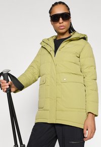 Gaiši zaļa puskosta jaka ar kapuci, pogām, divām priekšējām kabatām un teksturētu audumu. Modelis tur pārgājiena kāju, valkājot melnas saulesbrilles.