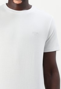 Jasnoszary t-shirt typu crew neck z krótkimi rękawami, z subtelnym logo "JOOP! JEANS" nadrukowanym w jasnoszarym kolorze na klatce piersiowej. Miękka tekstura.
