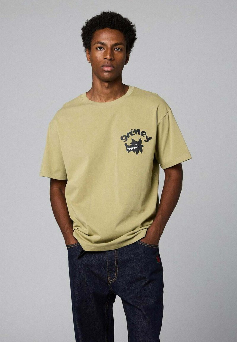 T-shirt in cotone verde oliva con un logo grafico nero raffigurante un mostro sul lato sinistro del petto. Fit rilassato, stile colletto rotondo.