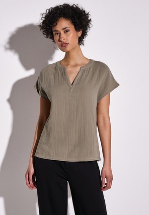 Blusa caqui con cuello, de manga corta, con tejido texturizado y escote en V. Combinada con pantalones de pierna ancha negros, que tienen un brillo sutil.