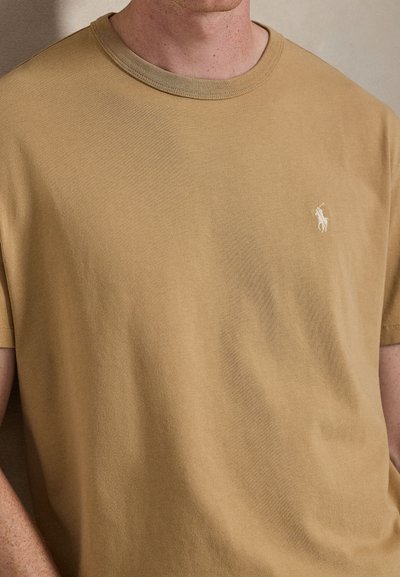 Tan cotton T-shirt with a round collar, featuring a small white logo on the left chest and short sleeves. Smooth, soft texture.