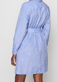 Blårandig klänning med långa ärmar, prydd med delikat guldbroderi, en midjebälte och knäppning baktill. Tillverkad av lättviktsmaterial.