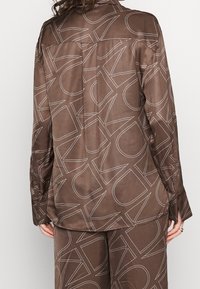 Harmaan satiinipaidat ja -housut, joissa on suuri, päällekkäinen valkoinen Calvin Klein -logo, esitetty takaa seisovalla henkilöllä.