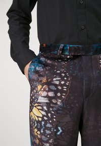 Multicolored patterned trousers feature a butterfly design against a dark background with a black shirt partially visible. Textured fabric.