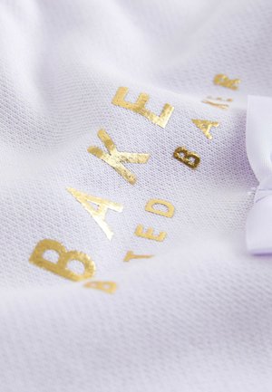 Hvidt stof med en tekstureret overflade prydet med guldtryk med teksten "BAKED BARI." En lys lilla båndaccent ses.