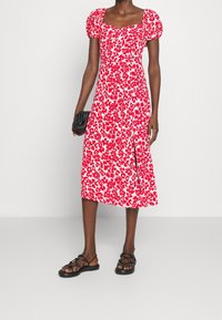 Rotes midi Kleid mit Blumenmuster, Puffärmeln, quadratischem Ausschnitt und einem seitlichen Schlitz. Kombiniert mit schwarzen Riemchensandalen und einer strukturierten schwarzen Clutch.