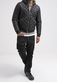 Czarna pikowana kurtka z kapturem, z przodem na zamek błyskawiczny i ściągaczami na rękawach, noszona na jasnoszarym swetrze i czarnych spodniach. Czarne buty.