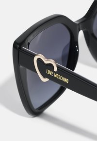 Črne sončne očala z drzno pravo, sivo lečo in zlatim poudarkom v obliki srca na črnem ročaju, na katerem je vgravirano "LOVE MOSCHINO."